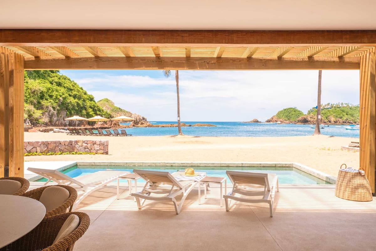 Zona de piscina con sillones de descanso y una mesa, con vista a una playa con sombrillas y sillones.