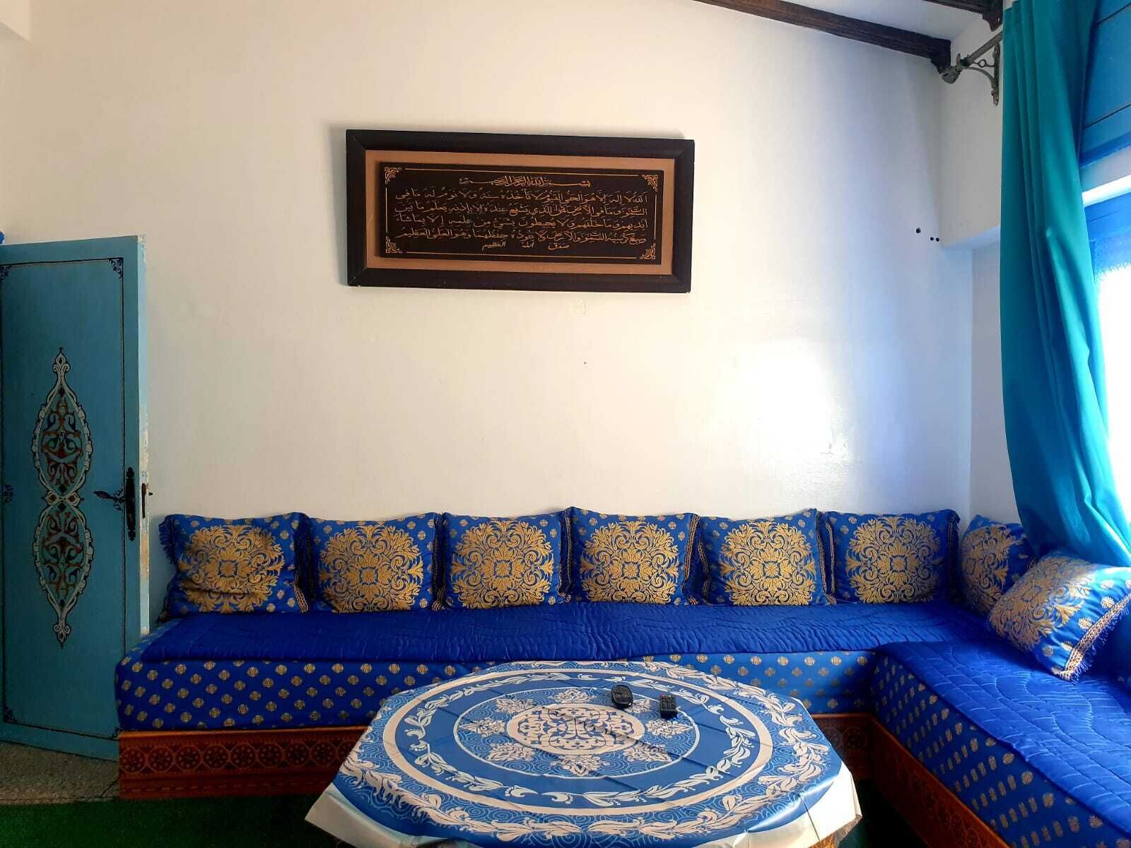 Una habitación con un sofá azul, un otoman con estampado, una obra de arte con caligrafía árabe enmarcada en la pared, y una puerta azul con diseños intrincados.
