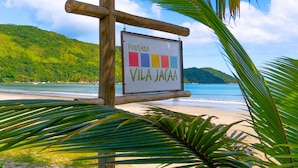 Ein hölzernes Schild mit der Aufschrift 'Pousada Vila Jacaa' vor einer Strandkulisse mit Palmen und blauem Himmel.