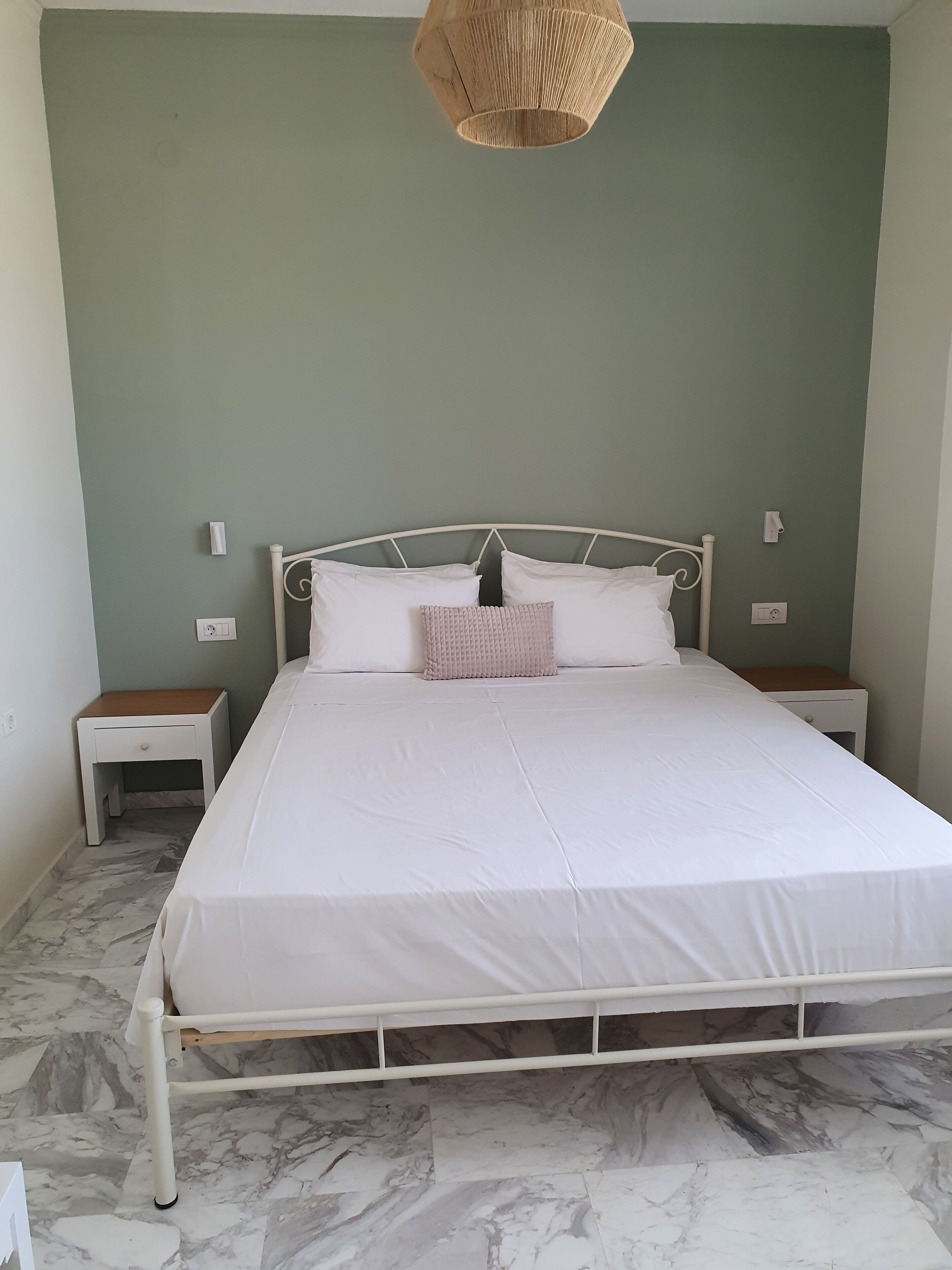 Ein ordentlich bezogenes Bett mit weißer Bettwäsche, einem weißen Metallgestell und einem gewobenen Kopfteil. Zwei Nachttische mit weißen Oberflächen, einer mit einer Schublade und der andere mit einem Regal. Der Raum hat eine grüne Wand und einen Boden mit Marmoroptik.