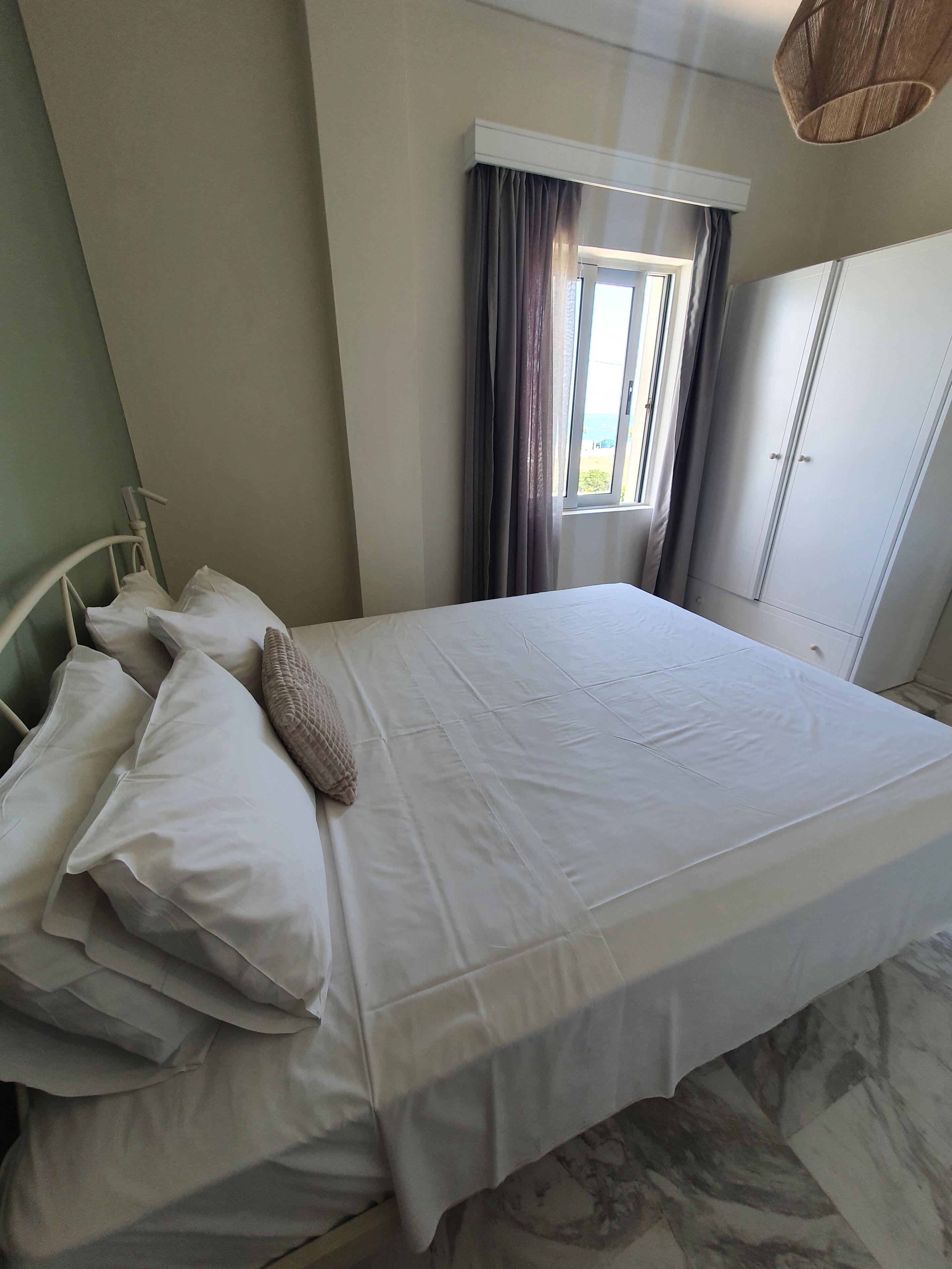 A neatly made bed with white linens and pillows in a room with a window, curtains, and a wardrobe.