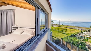 A bedroom with a large window overlooking a grassy area and the sea. - Oliva Beach Studios & Apartments (Platanias)