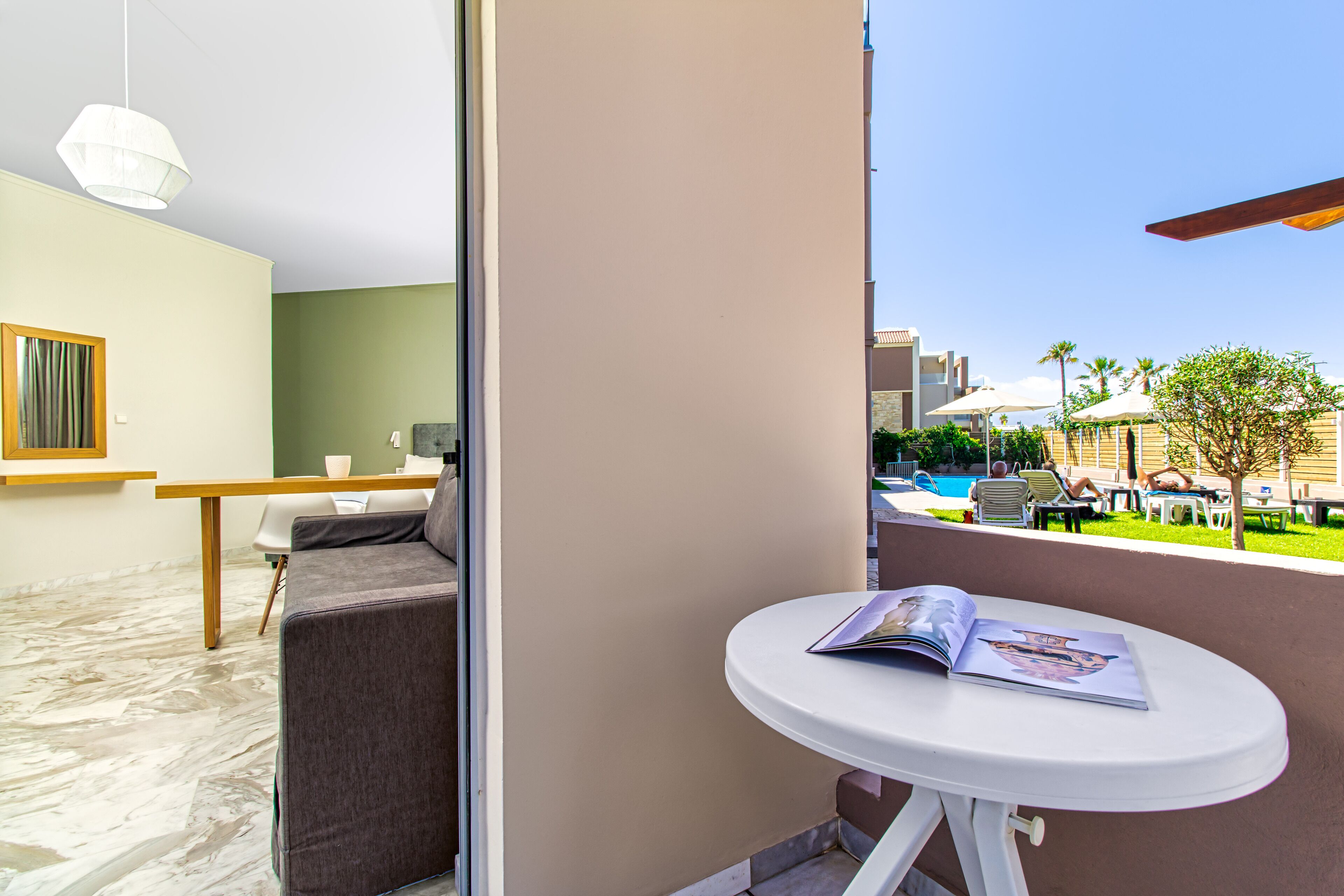 A balcony with a white table and a magazine, a grey sofa, and a view of a pool area with umbrellas and lounge chairs.