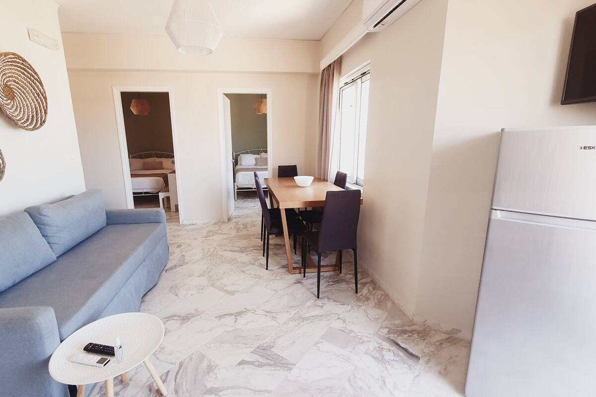 A modern living room with a sofa, a small table with a remote control, and a dining area with a table and chairs.