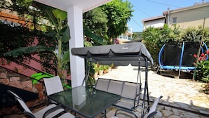 Eine Terrasse im Freien mit Glastisch und Stühlen, einem überdachten Sitzbereich und einem Trampolin.