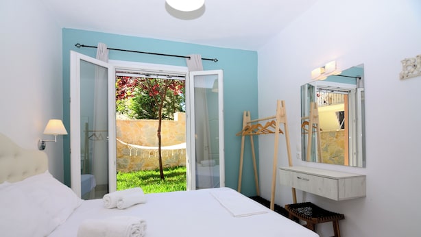 Ein Schlafzimmer mit Bett, Schreibtisch, Spiegel und Blick auf einen Garten durch eine offene Tür.