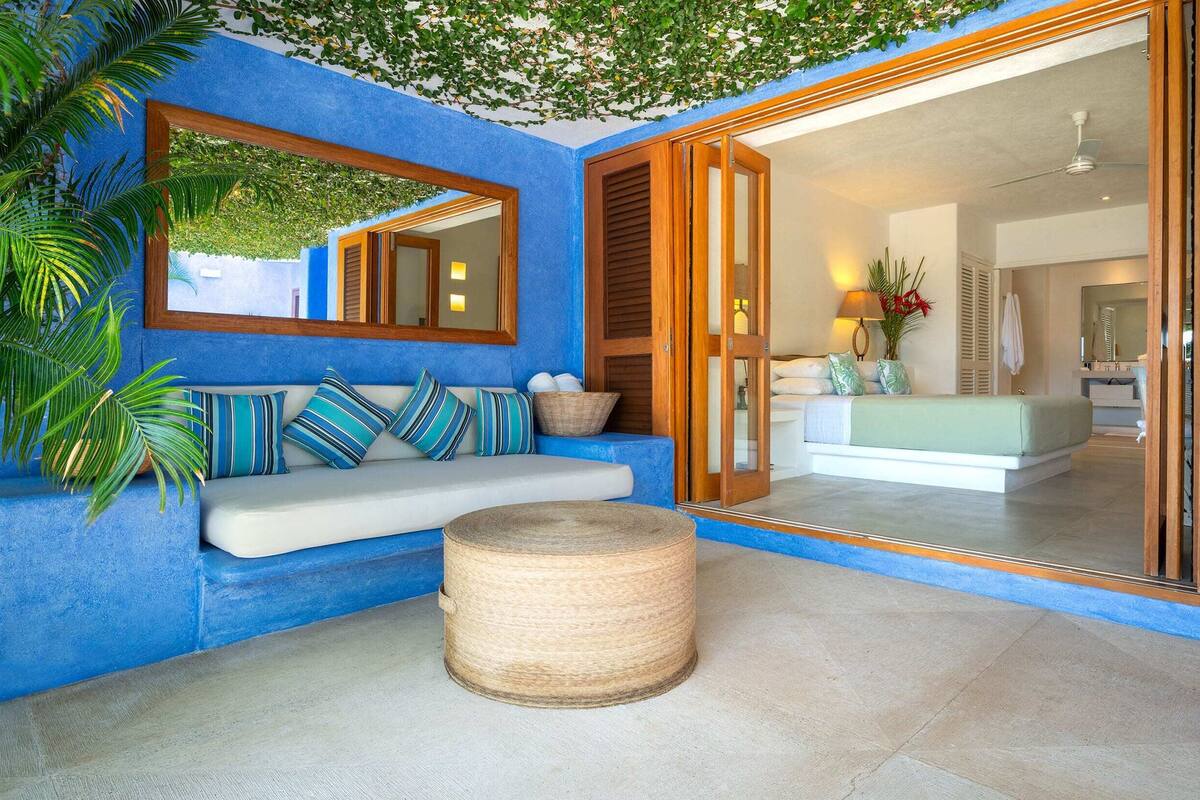 Ein Zimmer mit blauer Wand, einem spiegelartigen Wandbild mit Holzrahmen, einer Couch mit gestreiften Kissen, einem gewebten Hocker und einer Schiebetür zu einem Badezimmer.