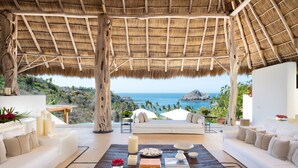 Amplia habitación con techo de paja, muebles blancos, una mesa de centro de madera y vista al océano.