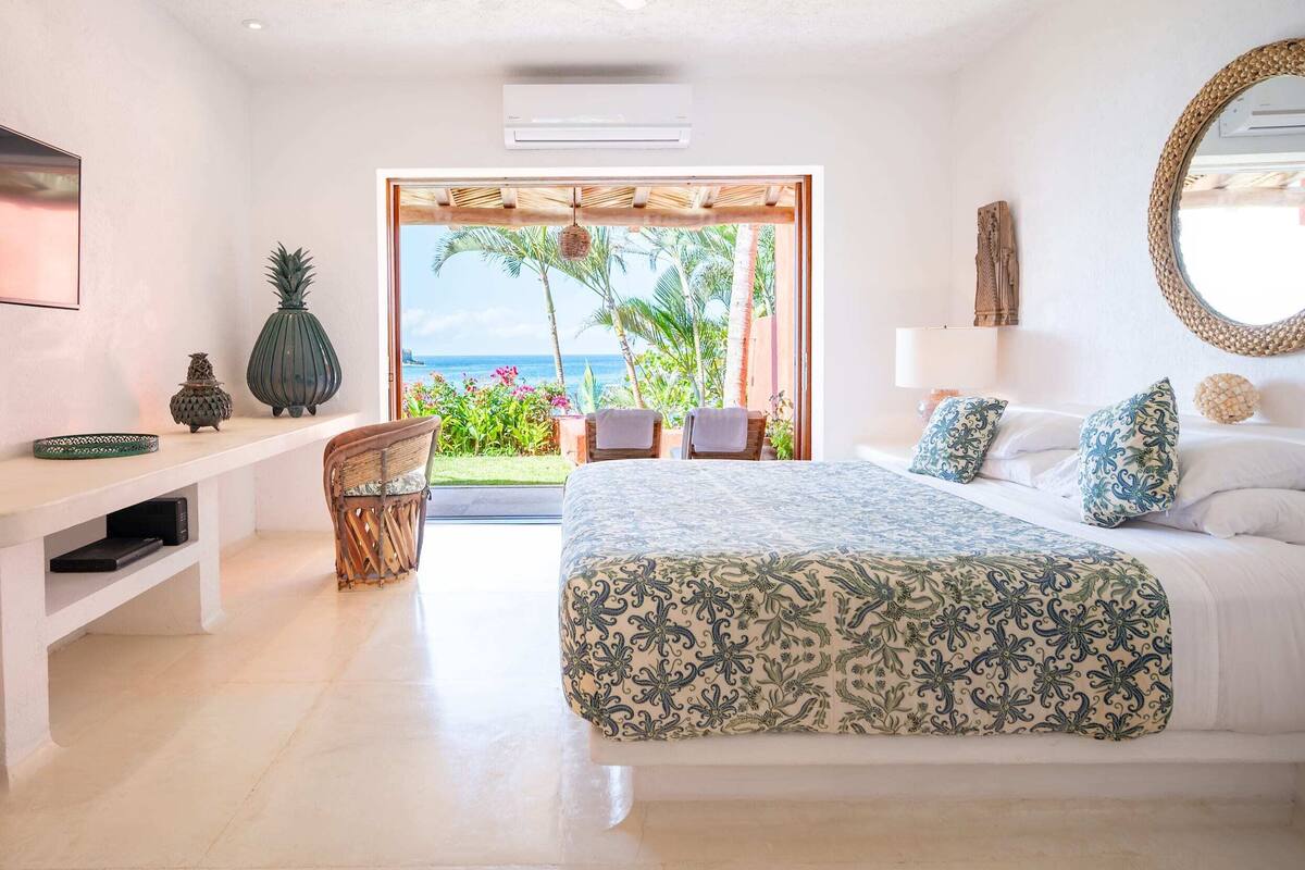 Ein Schlafzimmer mit Bett, einer Bank, einem Sessel, einem großen Fenster und Blick auf den Ozean.