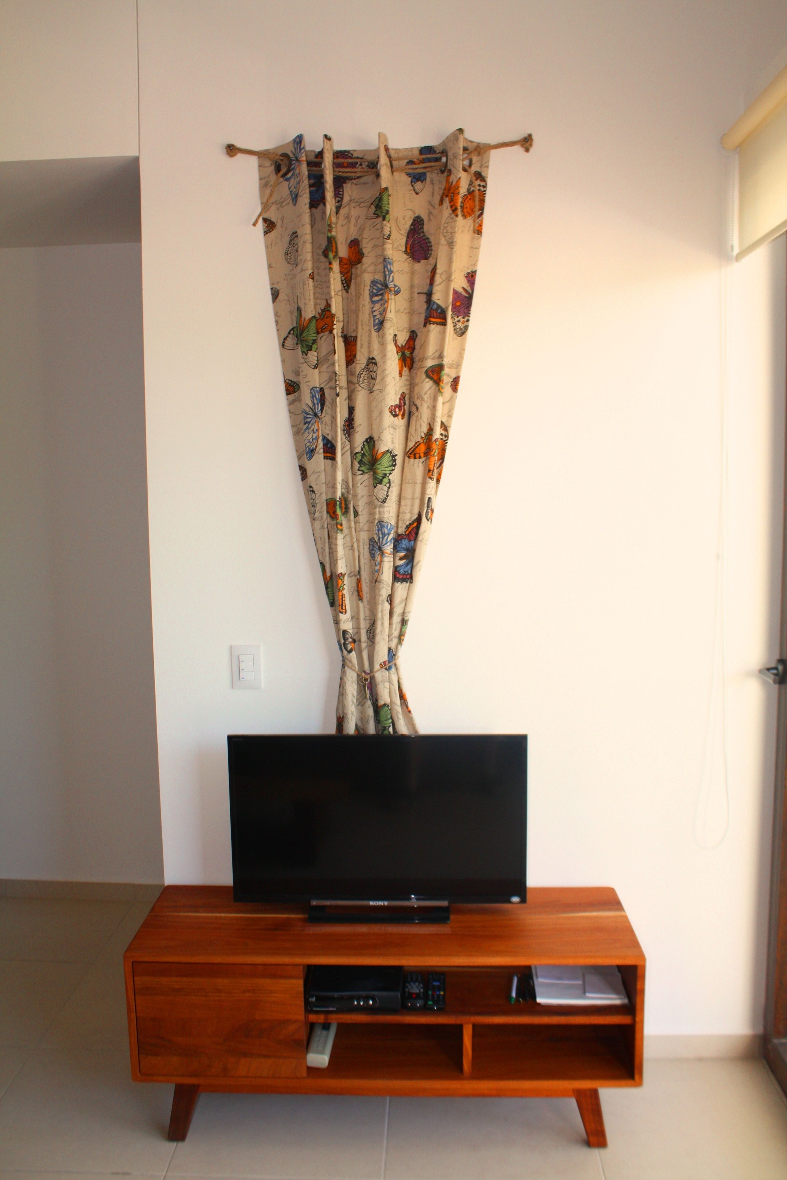 A wooden TV stand with a flat-screen TV and a DVD player, positioned against a white wall with a curtain featuring a butterfly pattern.