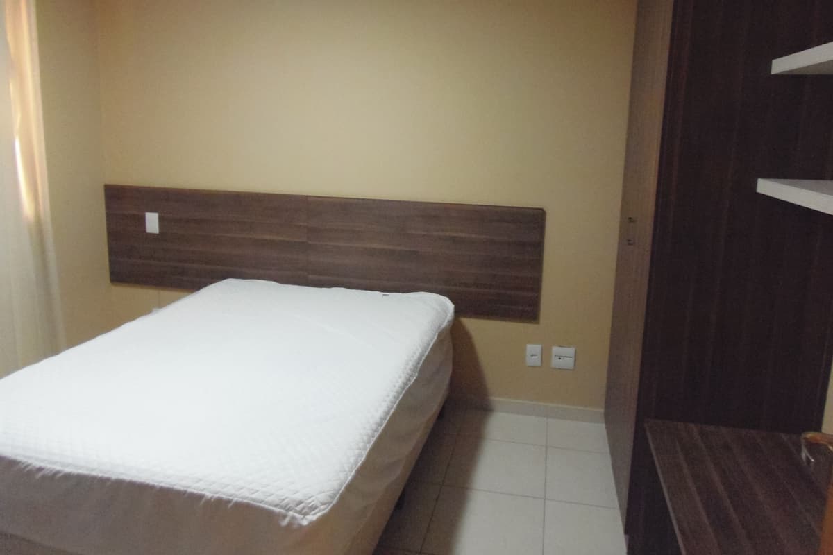 A bedroom with a bed, a wooden headboard, a small bedside table, and a wall-mounted shelf.