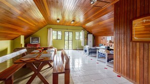 Um quarto espaçoso com teto alto, vigas de madeira, uma grande mesa de jantar com bancos, uma cama e um pequeno espaço de cozinha.