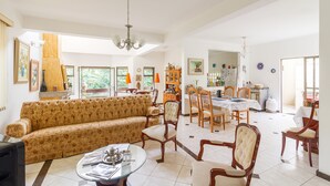 Ampla sala de estar com sofá, mesa de jantar e cadeiras, cozinha ao fundo e grandes janelas que permitem entrada de luz natural.