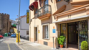 Hotel Dora, con un muro de piedra de fondo, un balcón con barandillas de hierro forjado y una pequeña mesa con plantas de maceta en el exterior.