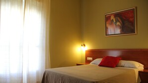 Una habitación de hotel con cama, mesita de noche, un cuadro en la pared y una ventana con cortinas.