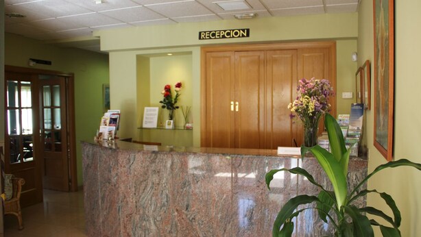 Un área de recepción con mostrador de mármol, una puerta de madera que dice “RECEPCIÓN” y una planta en maceta.