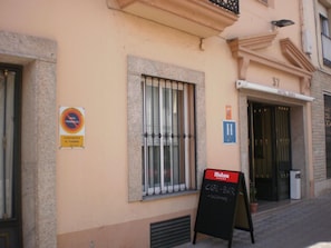 A building with a sign indicating a no parking zone, a window with metal bars, and a sign for a cafe-bar named Mahou. - Hotel Dora (Plasencia)