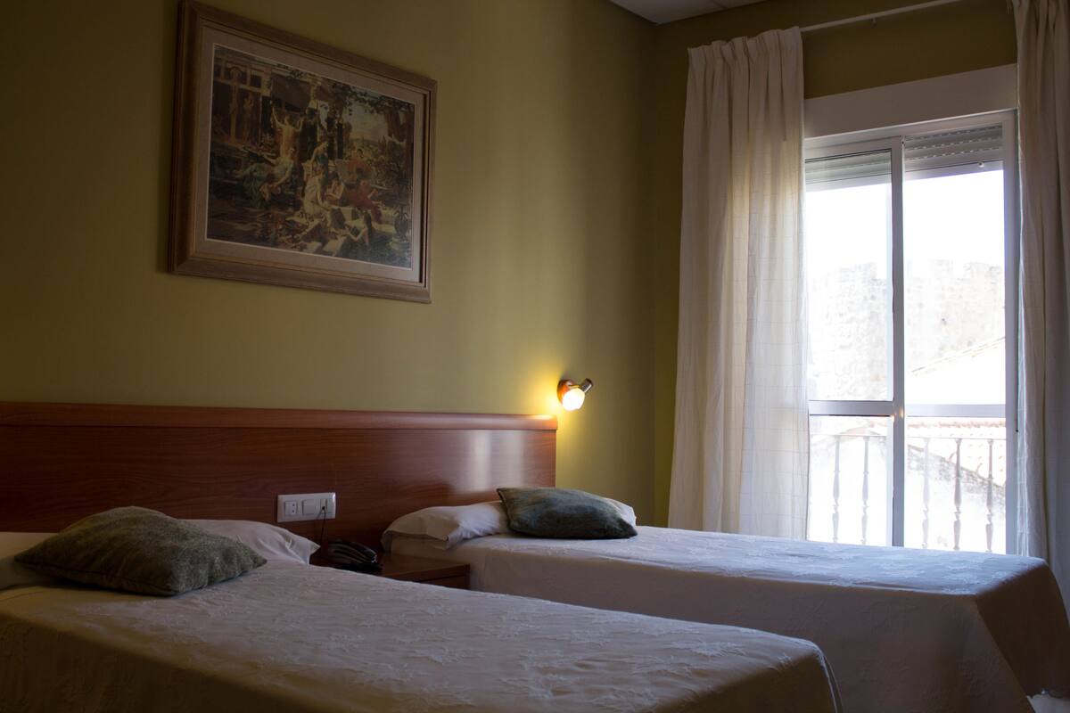 Quarto de hotel com duas camas, uma mesa de cabeceira, uma luminária na parede, um quadro e uma janela com cortinas.