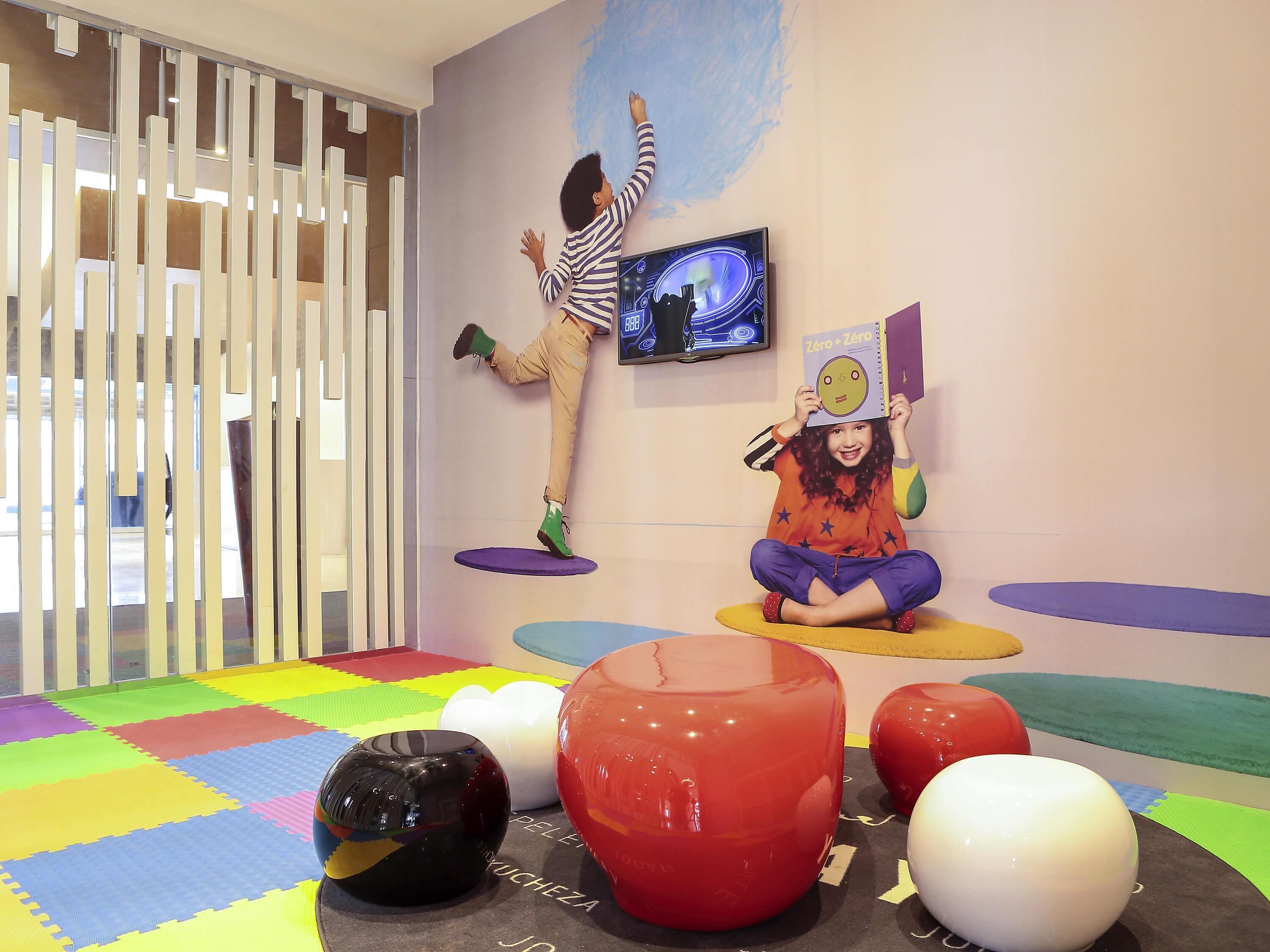 A room with colorful play mats, a child on a hanging platform, and a child sitting on a mat.