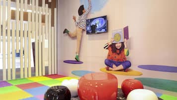 A room with colorful play mats, a child on a hanging platform, and a child sitting on a mat.