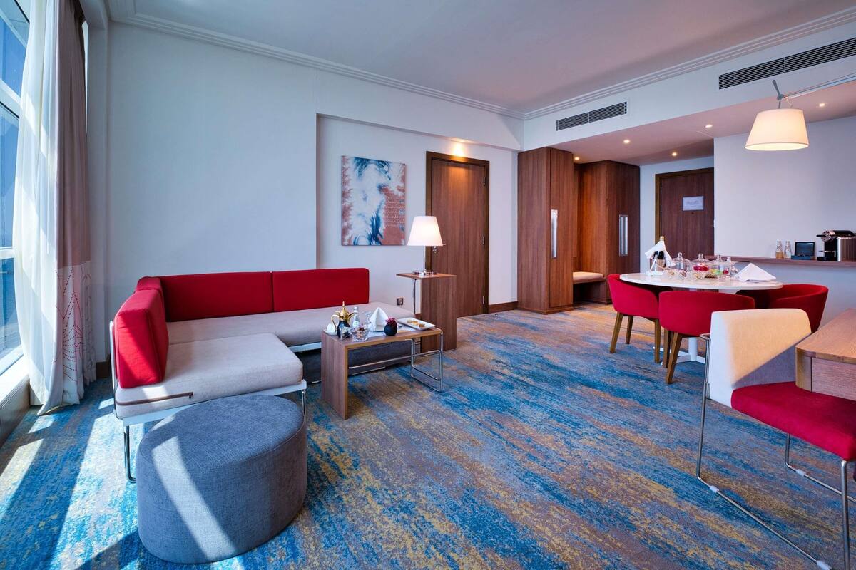 Una habitación de hotel moderna con un sofá rojo, un reposapiés gris, una mesa de comedor con sillas rojas y una zona de cocina.