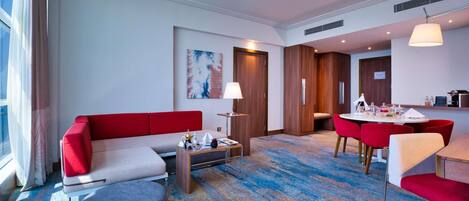 A modern hotel room with a red sofa, a grey ottoman, a dining table with red chairs, and a kitchen area.