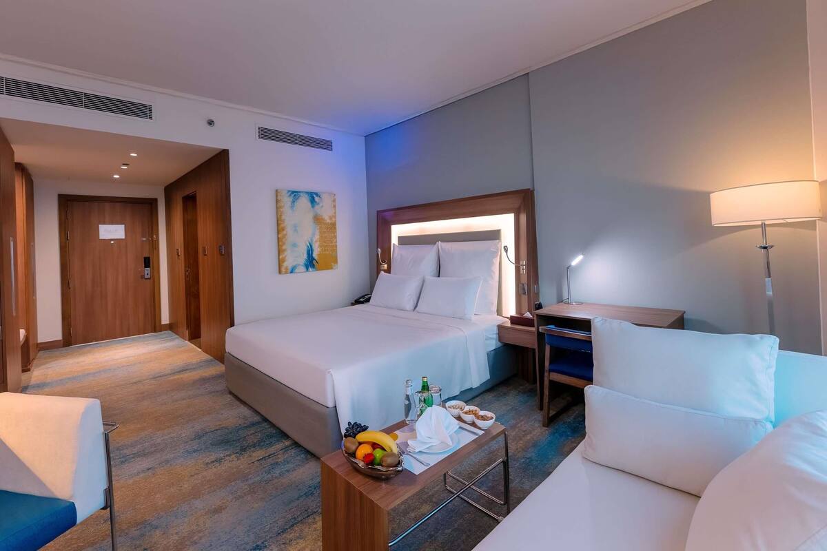 Habitación de hotel con cama, escritorio, silla, mesita de noche con fruta y lámpara.