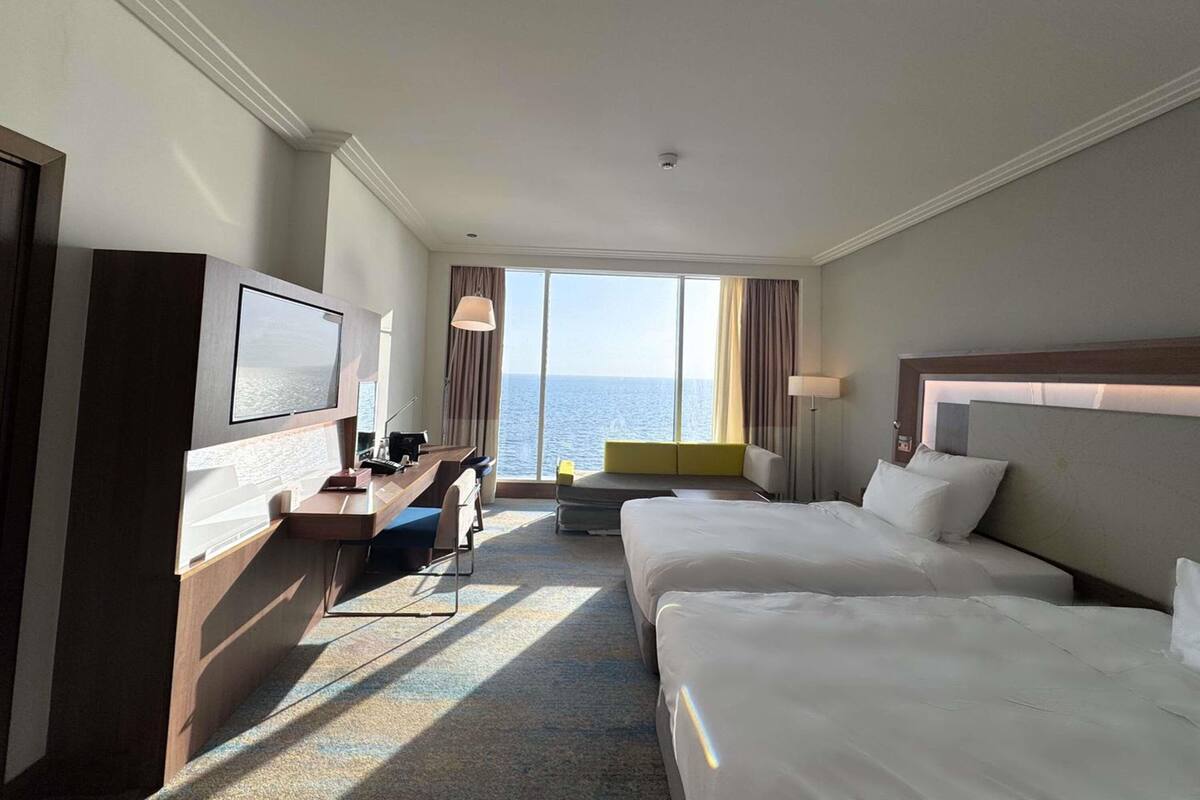 Habitación de hotel con cama, escritorio, silla, televisor, ventana con vista al mar y lámpara.