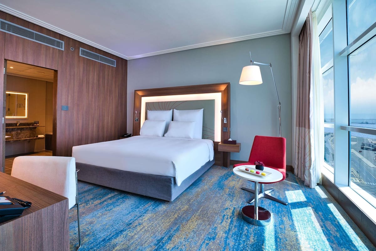 A hotel room with a large bed, a desk, a chair, a small table with a lamp, and a view of the city through large windows.