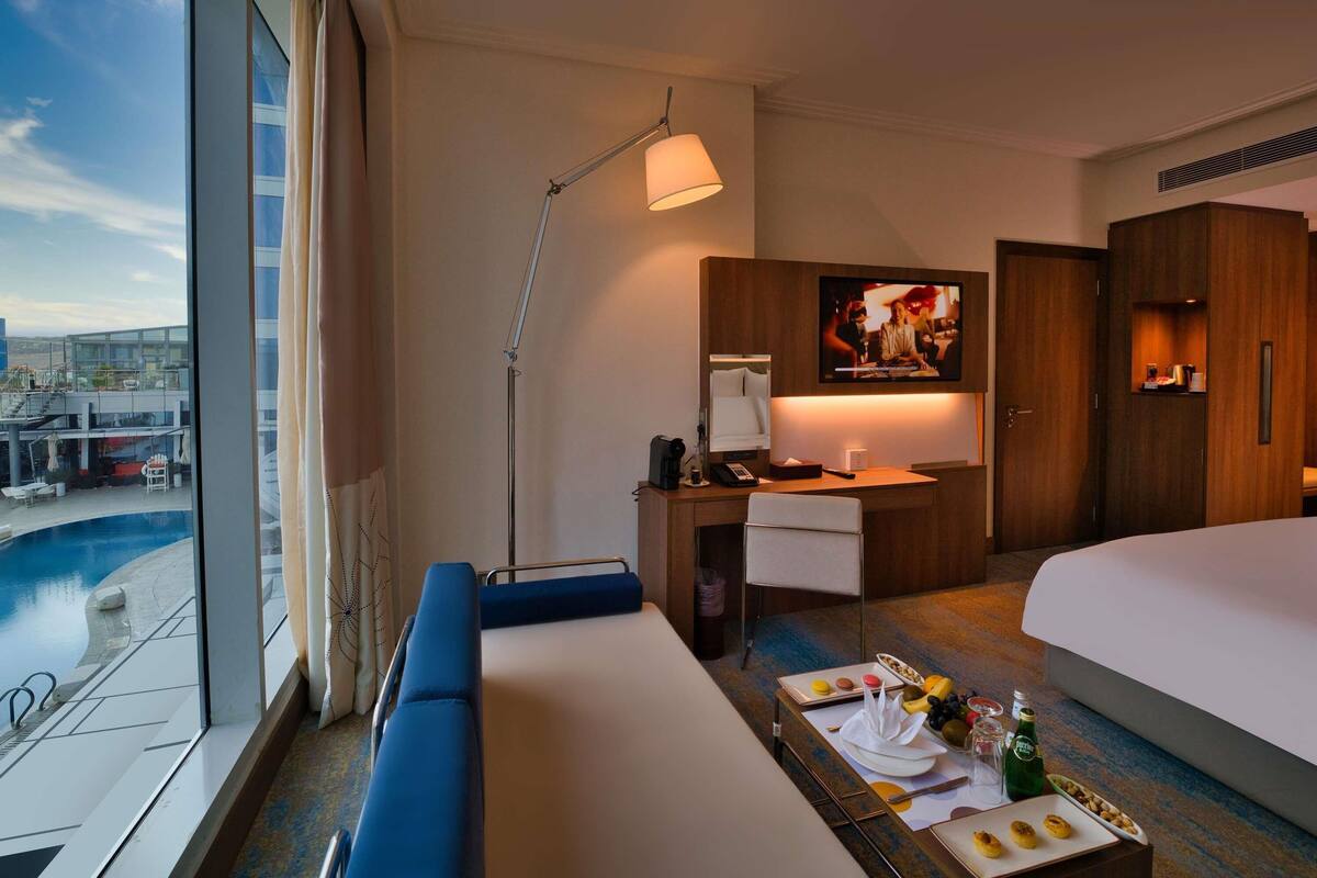 Habitación de hotel con una cama grande, un escritorio con televisor, un sofá, un rincón comedor con desayuno y vistas a una piscina y edificios.