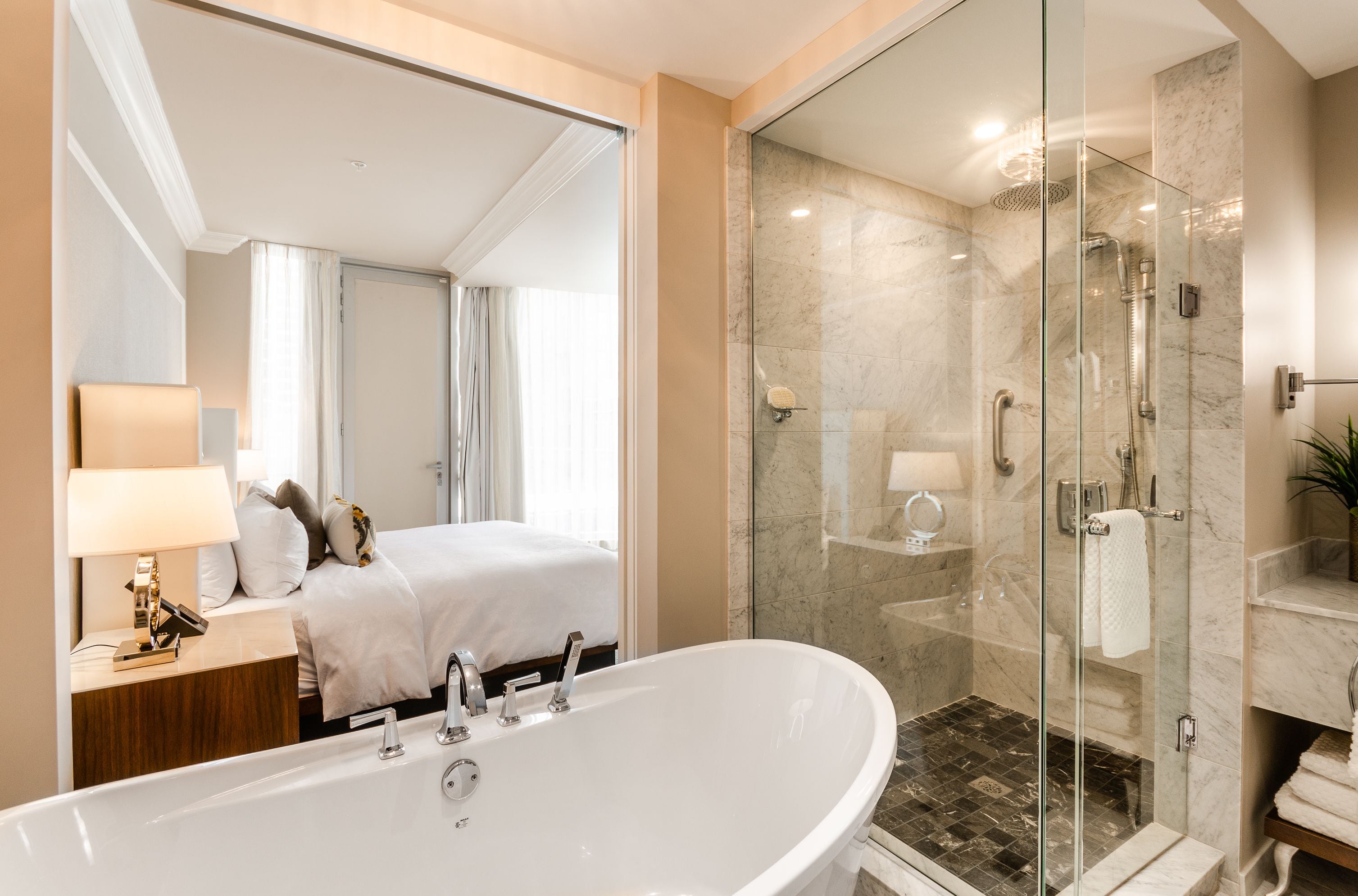 Un baño moderno con una bañera independiente grande, una ducha con cerramiento de vidrio, y una cama a la vista con mesita de noche y lámpara.