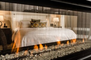 A hotel room with a bed, pillows, and a fireplace with burning logs.