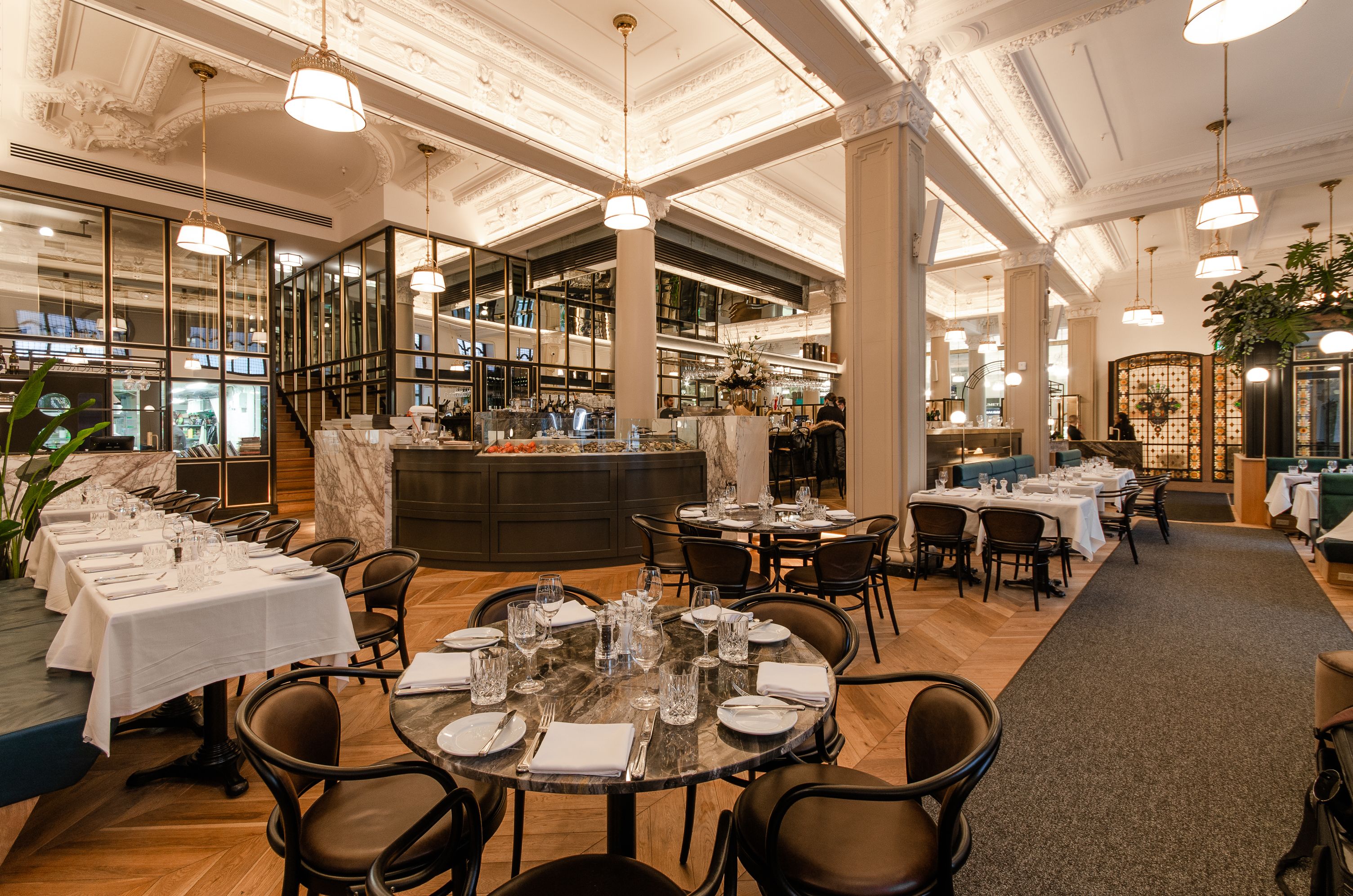Ampla área de refeições de hotel, com várias mesas dispostas para hóspedes, um bar central e uma planta decorativa.