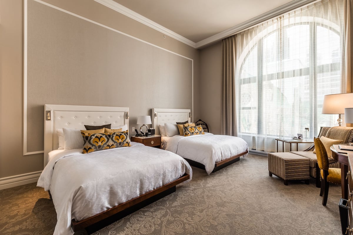 Ein Hotelzimmer mit zwei Betten, einem Schreibtisch, einem Stuhl und einem großen Fenster mit Vorhängen.