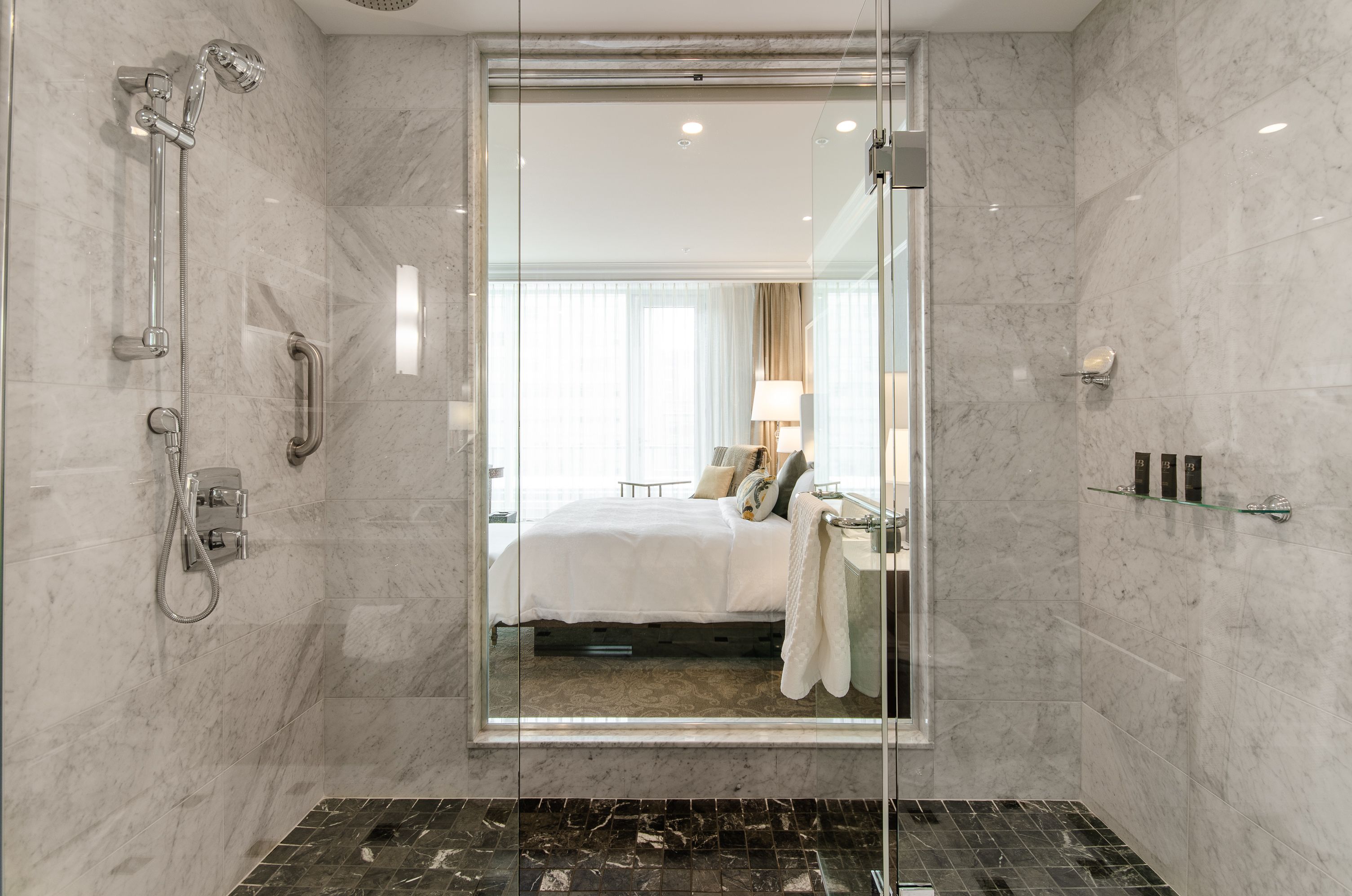 A modern bathroom with a glass-enclosed shower, marble walls, and a visible bedroom with a bed and window.