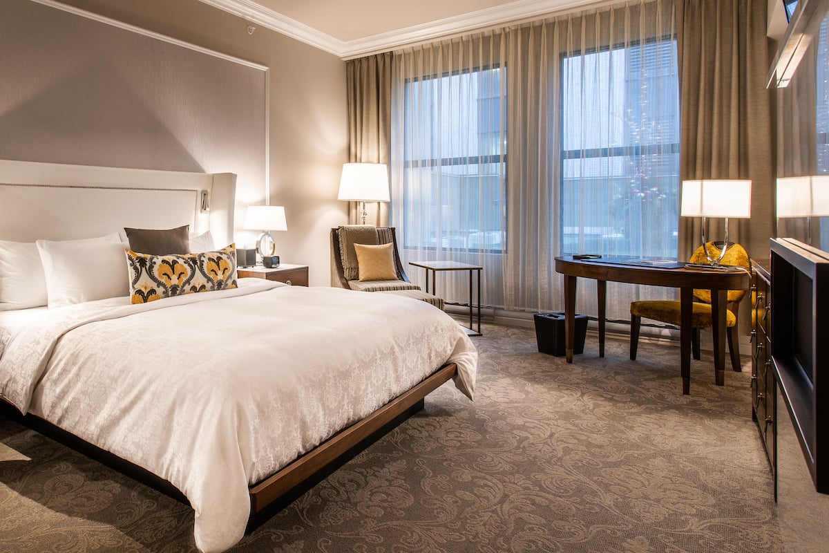 Ein Hotelzimmer mit einem großen Bett, zwei Nachttischen mit Lampen, einem Sessel, einem Schreibtisch und einem Fernseher.
