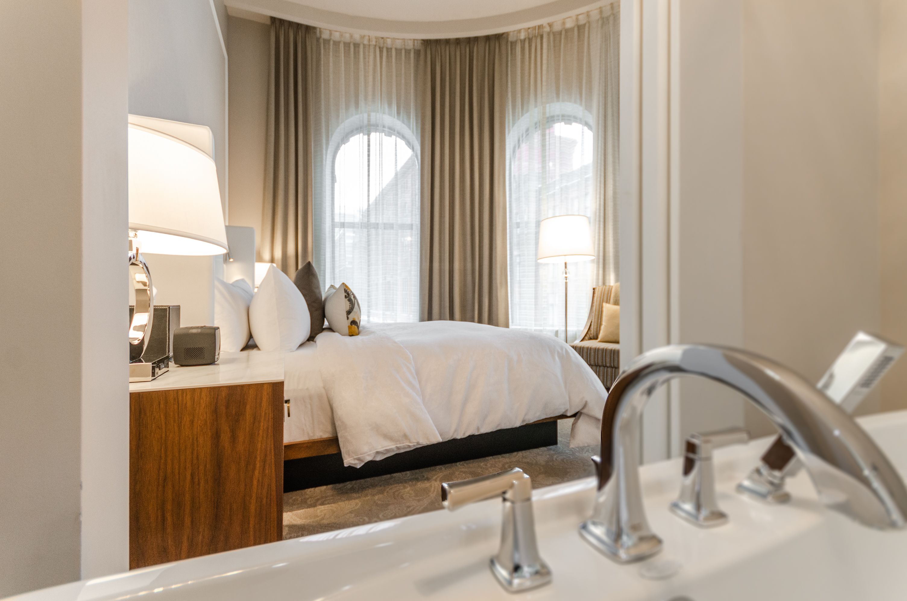 A hotel room with a bed, bedside lamps, a wooden nightstand, a large window with curtains, and a modern bathroom with a sink and mirror.