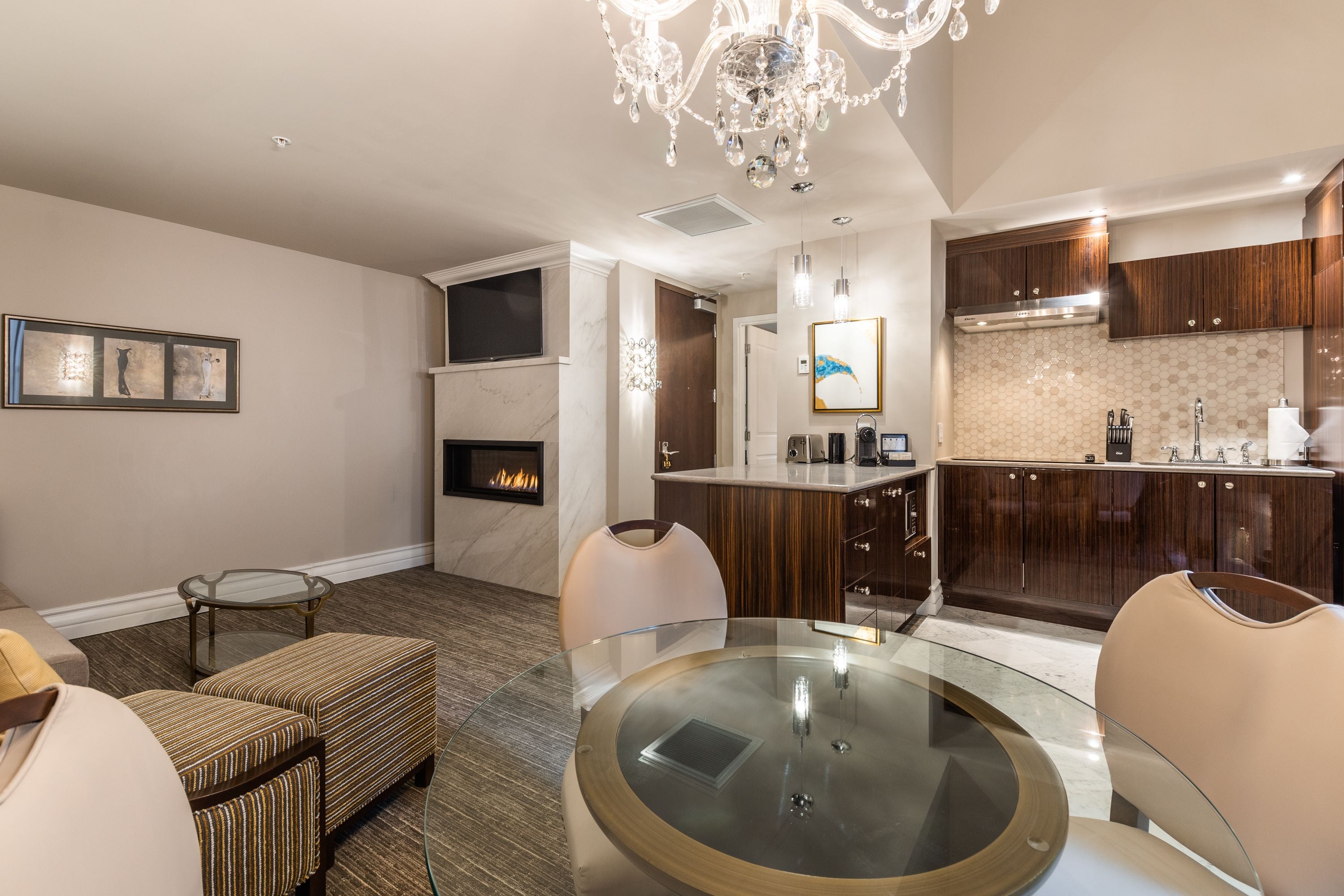 A modern living room with a glass dining table, comfortable seating, a fireplace, and a kitchen area with wooden cabinets and a marble backsplash.