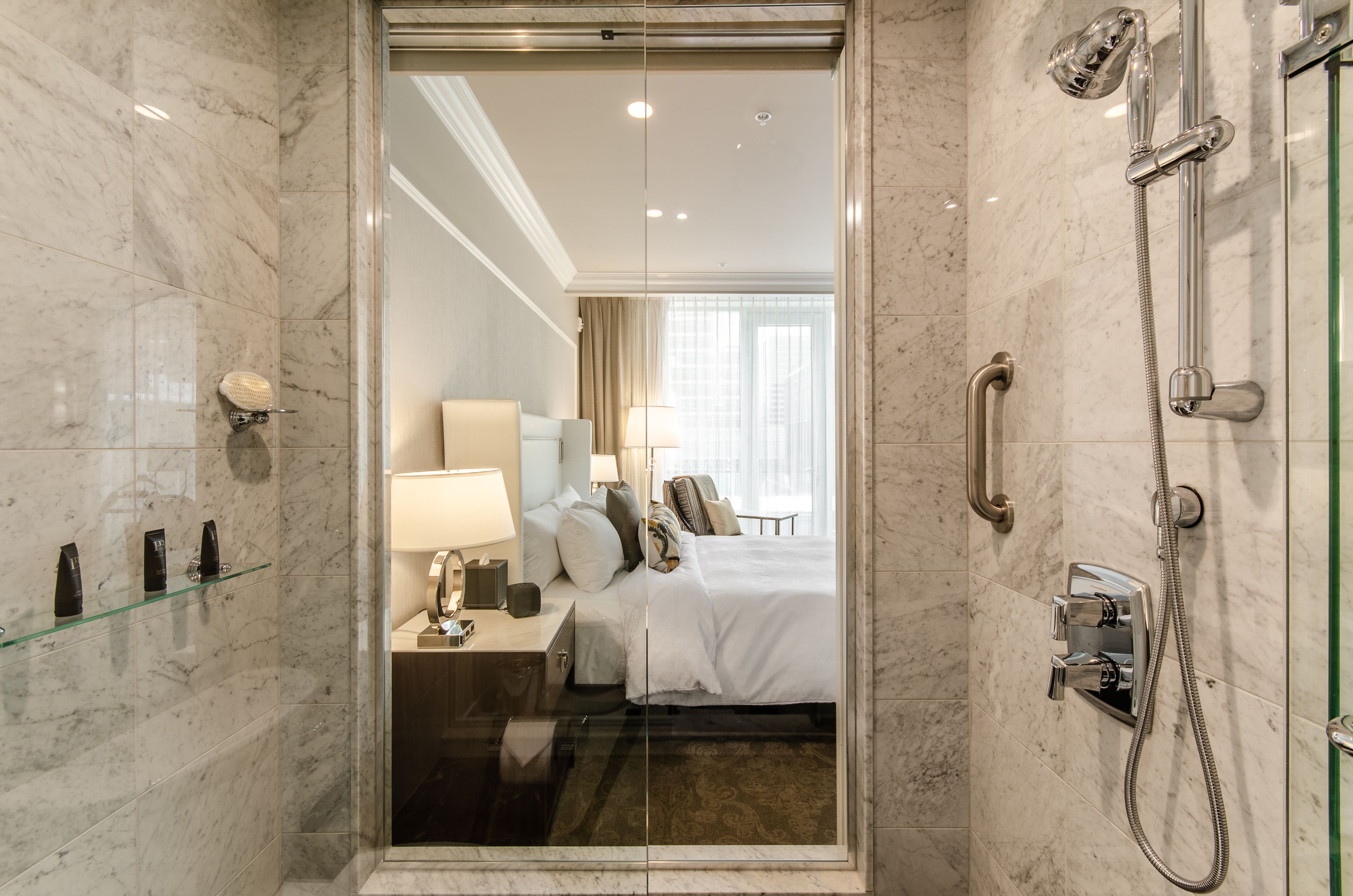 Un baño de hotel con cabina de ducha de vidrio, paredes de mármol, una cama con ropa blanca y una mesita de noche con lámpara.