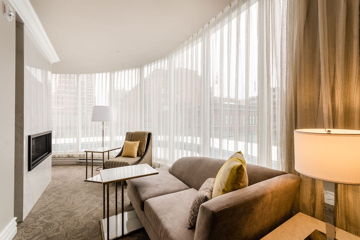 Ein modernes Hotelzimmer mit einer gebogenen Couch, einem kleinen Tisch, einem Stuhl, einem an der Wand montierten Fernseher und transparenten Vorhängen, die natürliches Licht hereinlassen.