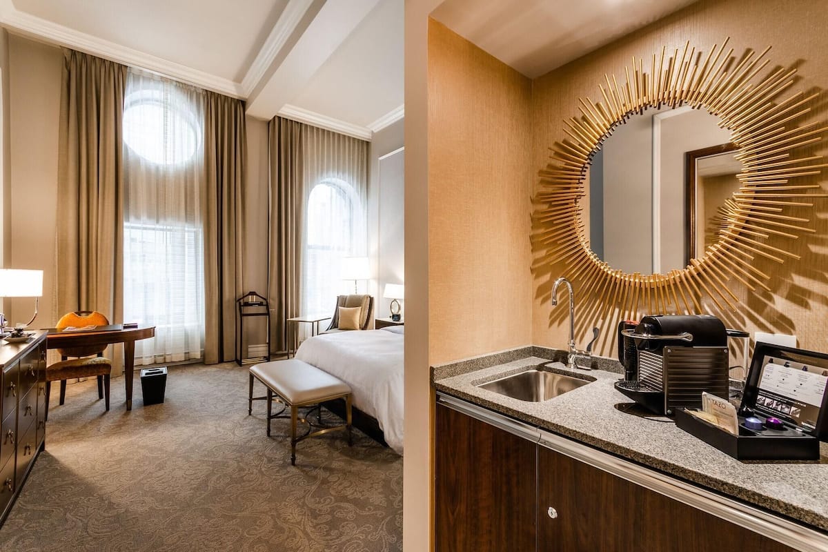 Ein Hotelzimmer mit Bett, Schreibtisch, Stuhl, Bank, großem Spiegel, Waschbecken und Kaffeemaschine.