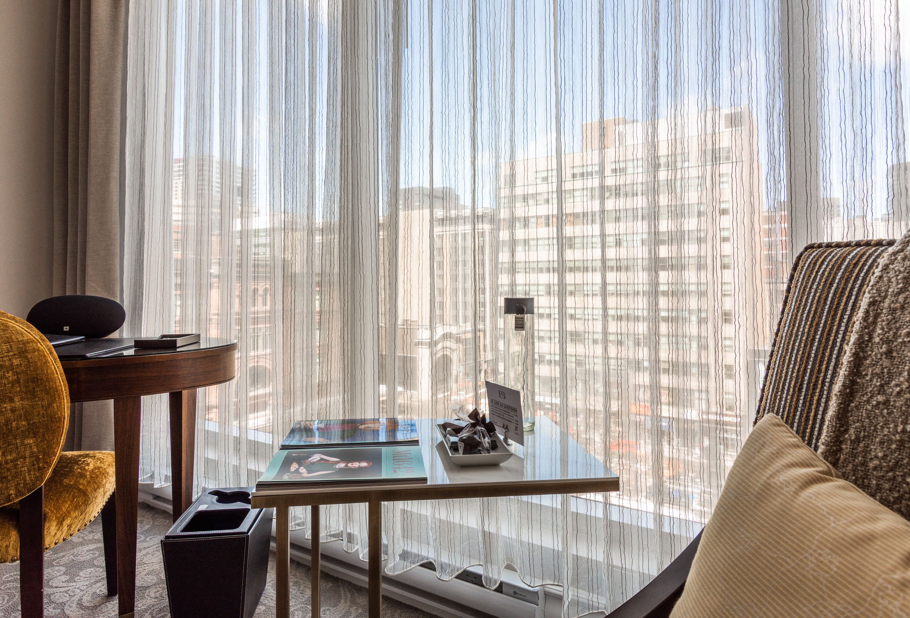 A hotel room with a view of a cityscape through sheer curtains, a round table with a laptop, a small trash bin, and a yellow armchair.