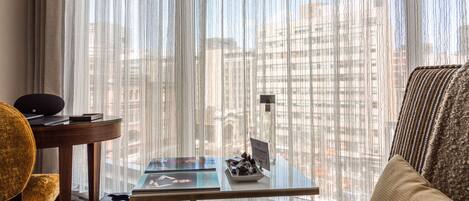 A hotel room with a view of a cityscape through sheer curtains, a round table with a laptop, a small trash bin, and a yellow armchair.