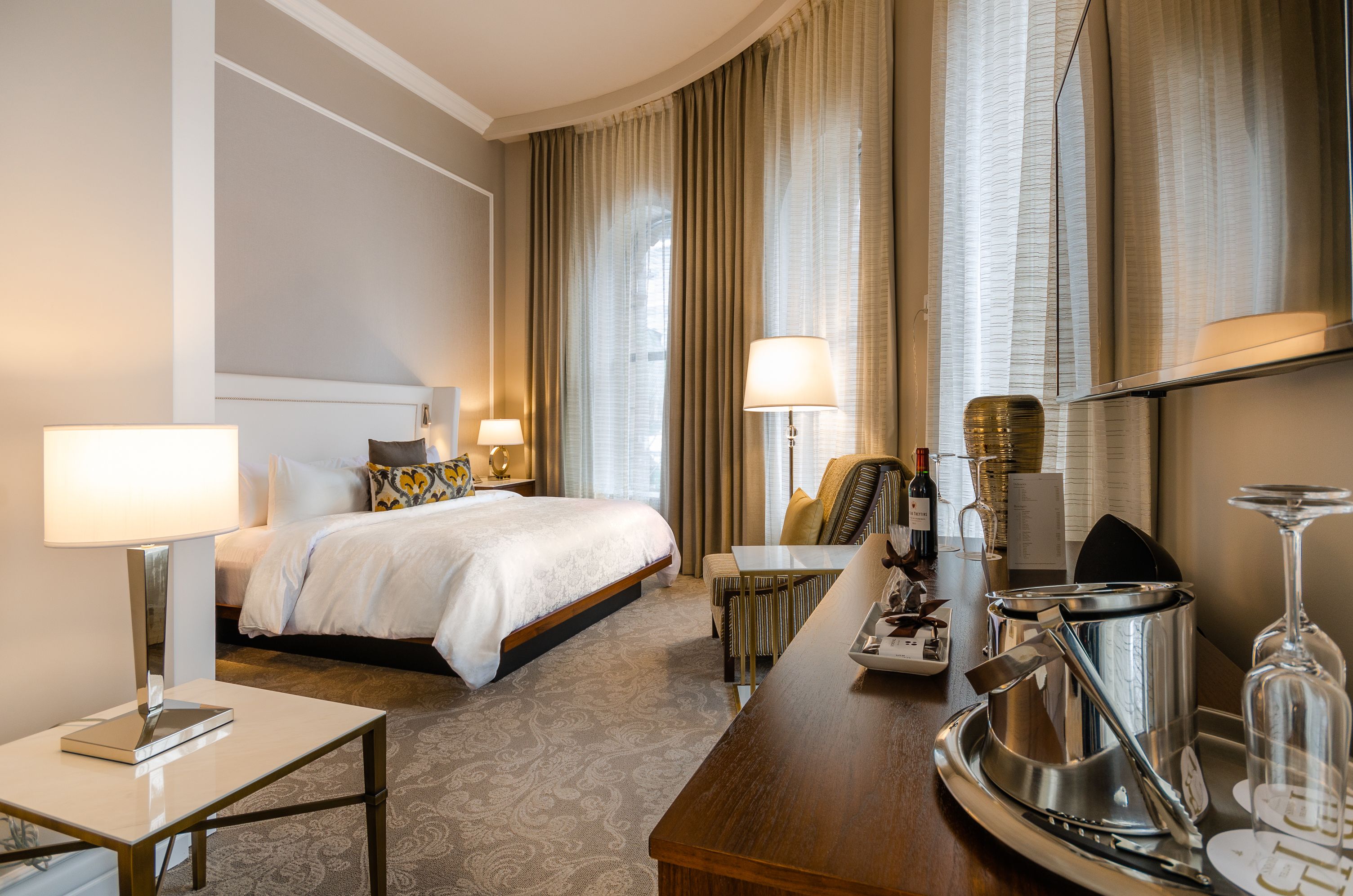 A hotel room with a bed, bedside tables, a desk, a chair, a minibar, and curtains.