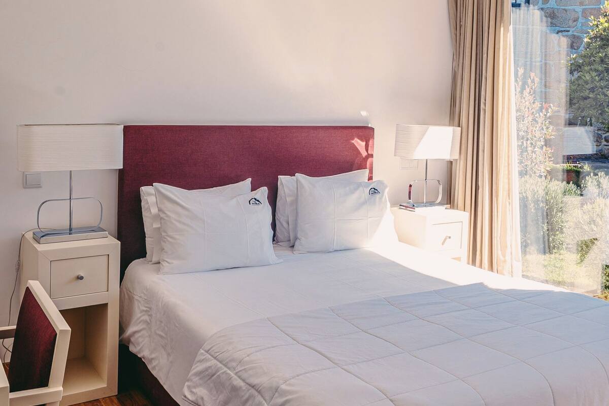 Una habitación de hotel con una cama grande, una mesita de noche, una silla y un ventanal con cortinas.