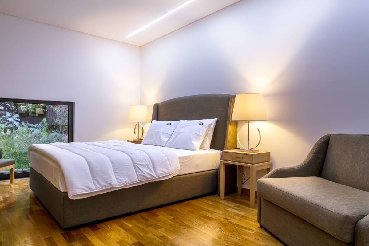 Una habitación de hotel moderna con una cama, dos mesitas de noche, una silla y un gran ventanal con vistas a zonas verdes.