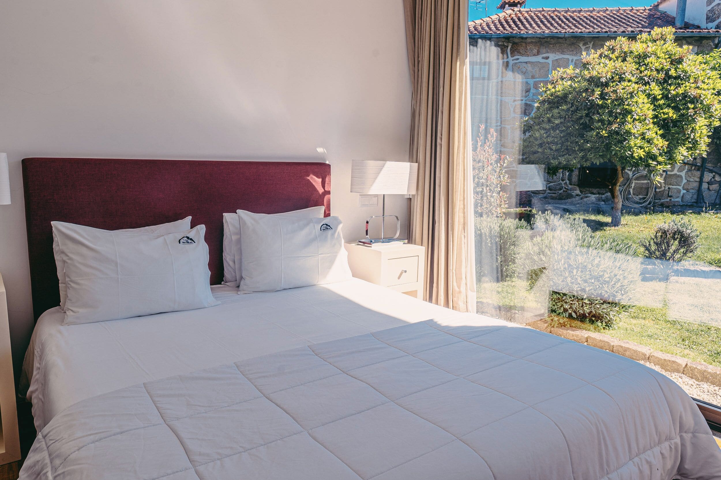 Habitación de hotel con una cama grande, una mesita de noche, una lámpara y vista a un patio con un árbol.