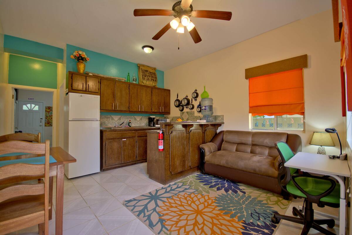 A compact living space with a kitchenette, a brown leather sofa, a dining table with chairs, and a patterned rug.