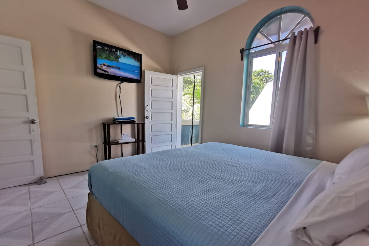 A bedroom with a bed, a TV mounted on the wall, a door, and a window with curtains.
