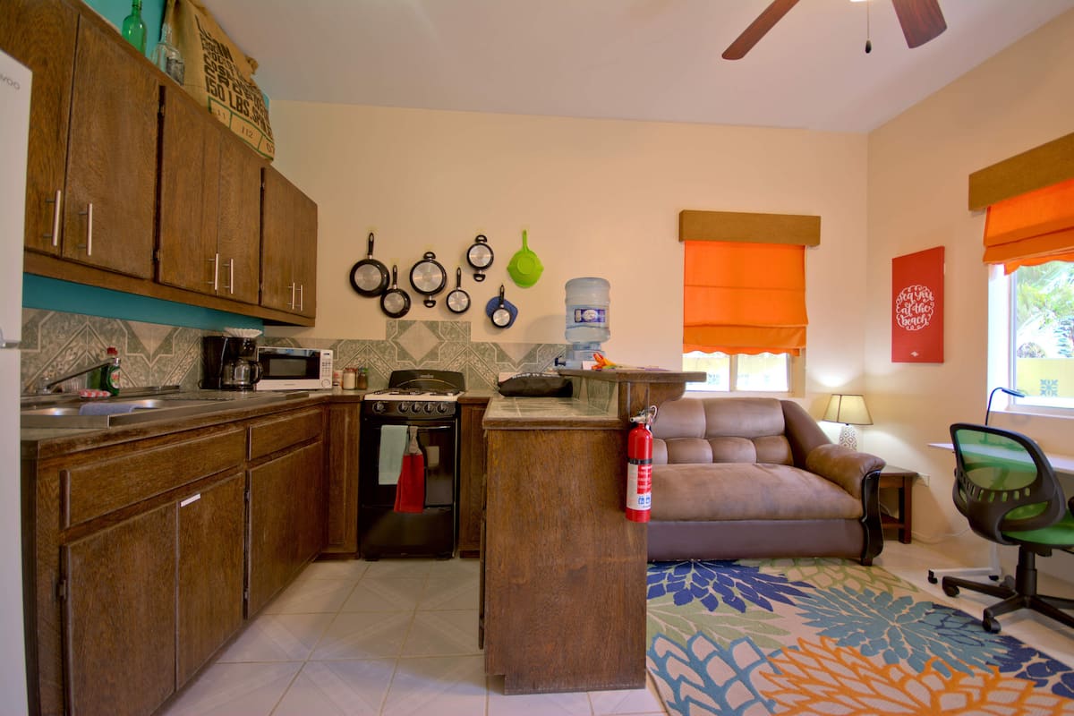 A kitchen with wooden cabinets, a microwave, stove, and a water dispenser. There is a sofa and a chair in the living area.
